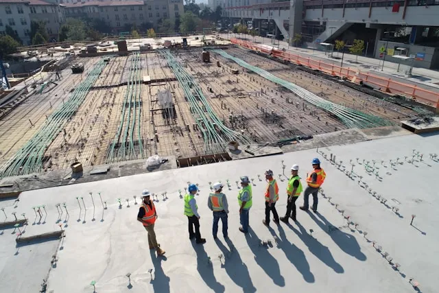 Construction workers in hard hats and high-visibility vests standing on a concrete deck beside a reinforced foundation